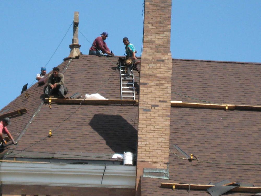 Roofers fitting new shingles on a pitched house roof beside a brick chimney